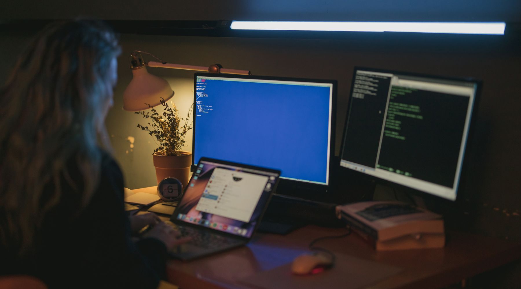A person working at a laptop and two monitors in a dark room
